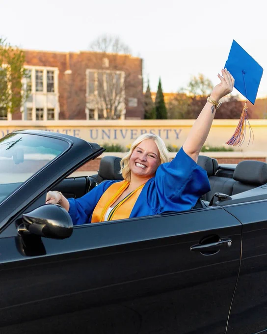 Boise State undergraduate student wearing graduation regalia in a car in front of the B Plaza.