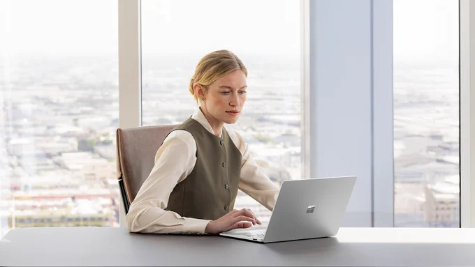 Business person sitting at a desk in front of a large window working on a Surface Laptop