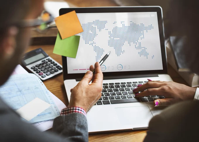 Stock image of a laptop screen with the map of the world. Two people can be seen looking at the screen. There are sticky notes on the laptop screen and papers and a calculator on the desk next to the laptop.