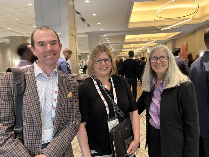 Three attendees smiling at a conference event. The person on the left is wearing a checkered blazer with a backpack, the middle person has a badge and folder, and the person on the right is in a black blazer. They are indoors with other attendees in the background.