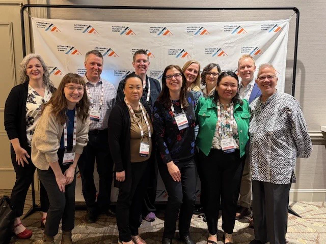 Group of 11 attendees at a conference standing in front of a 'step and repeat' backdrop printed with Affordable Housing Summit logos, smiling for a photo.