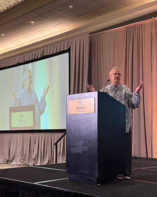 A person speaking at a podium with the Hilton logo, with a projected image of themselves in the background.