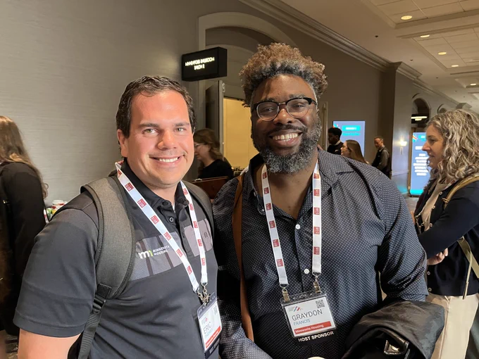 Two individuals at a conference, smiling and wearing lanyards with name tags. They're both wearing dark gray shirts.