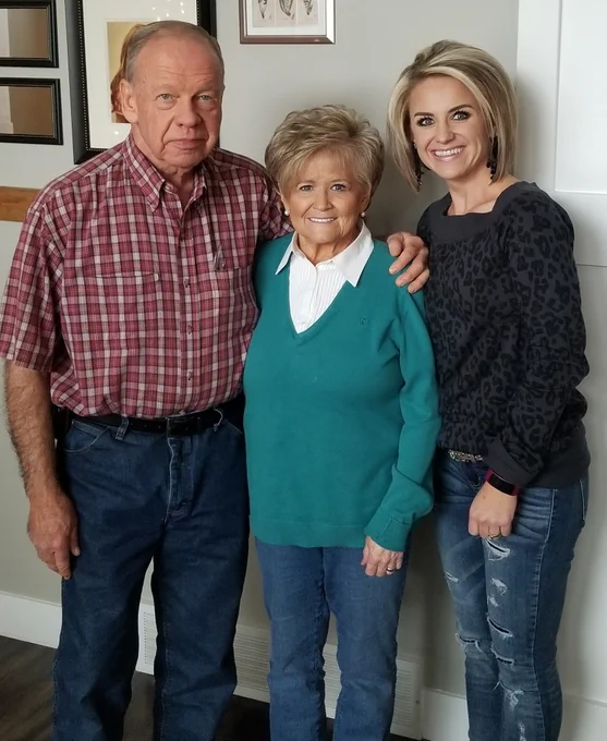 Gina Corry, wearing a black blouse and denim jeans, smiles and poses for a photo with both her father and mother.
