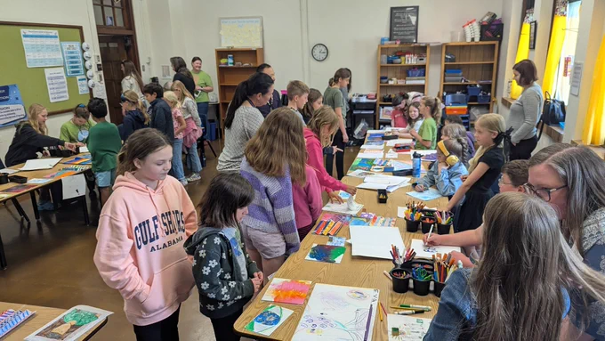 People walking around the classroom looking at student artwork displayed on tables