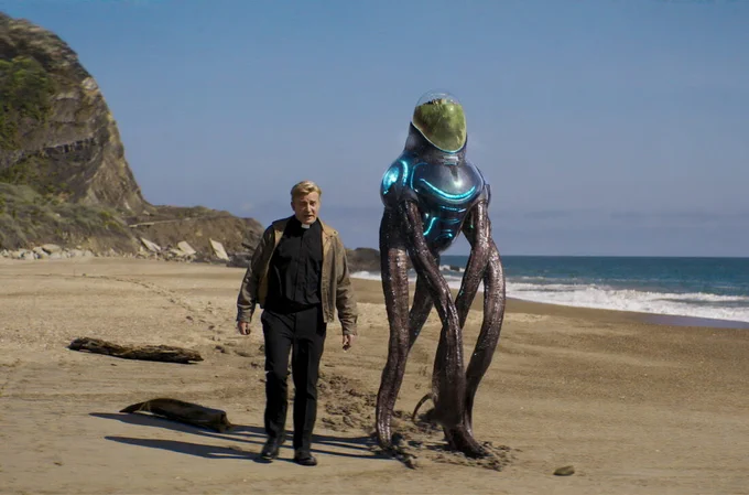 Rhys Darby in a priest outfit walking next to an alien on a beach