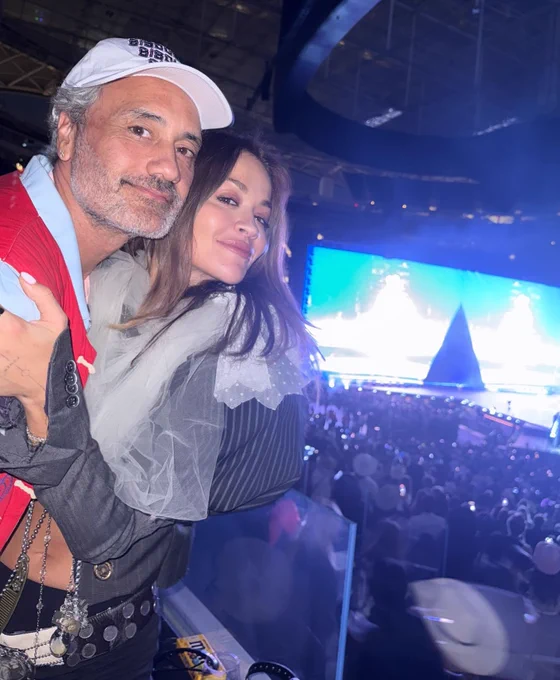 Taika Waititi and Rita Ora in a booth overlooking a stage.