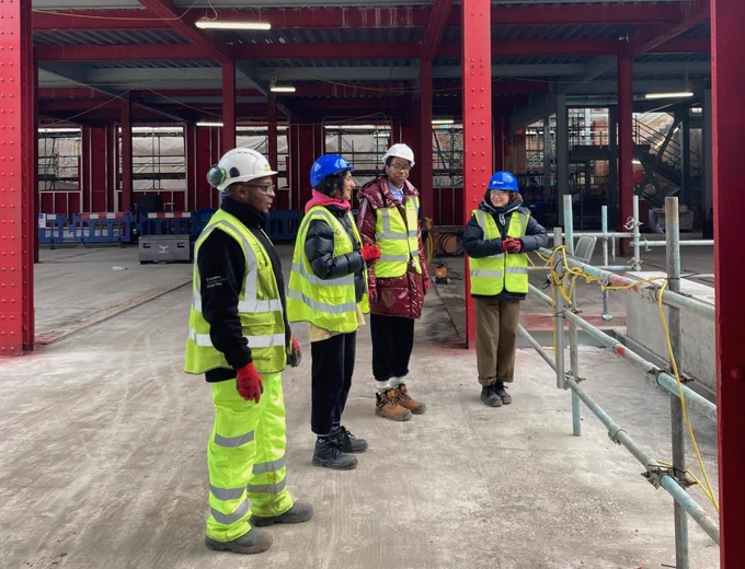 A landscape image of figures in personal protective equipment, including hard hats, eye goggles, and high-vis jackets. They stand within a building site, where red steel beams and concrete floors are visible. Image courtesy of Grand Union.