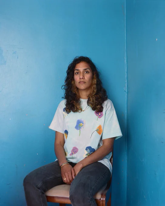 A portrait image of a person sitting on a wooden chair against a bright blue wall. The person looks directly to the camera, their long dark hair falling in waves around their shoulders. They wear dark jeans and a pale t-shirt covered in large images of different coloured poppies. Image by Robin Christian.