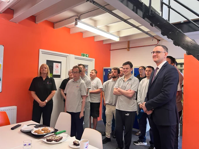 Ian is standing with a group of young men who are all wearing grey polo shirts with the Switch Now logo on them. In this image they are observing the VE Day 2 minute silence, and they are looking up at a screen watching the ceremony. They are inside a building where a table has plates of cakes in front of them.