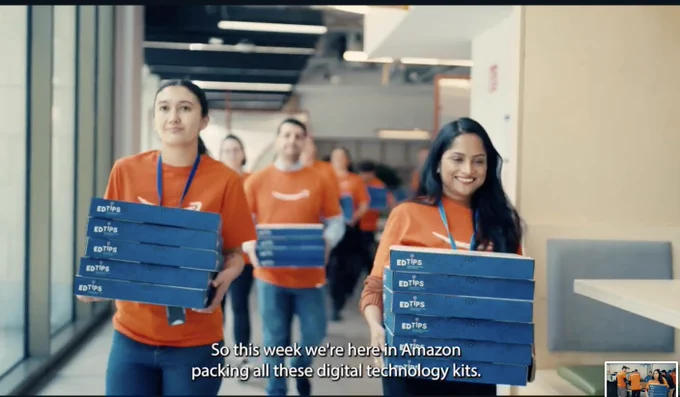 Amazon in Communities volunteers in orange shirts carry stacked 'EDTIPS' boxes down a hallway, preparing digital technology kits as part of an initiative to provide every primary school across Ireland with digital technology kits. 