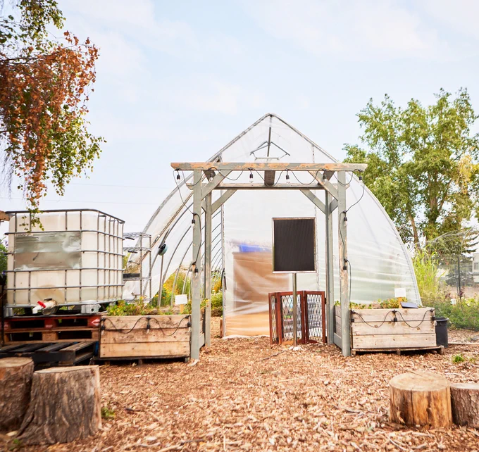 A greenhouse decorated with a wooden garden structure and raised garden beds on each side of the entrance of the greenhouse. Four logs are on both sides of the greenhouse. 