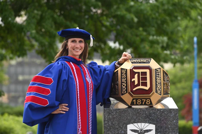 A Duquesne University senior in graduation regalia stands next to the Red Ring statue.