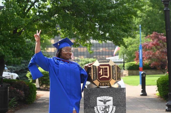 A Duquesne University senior in graduation regalia stands next to the Red Ring statue.