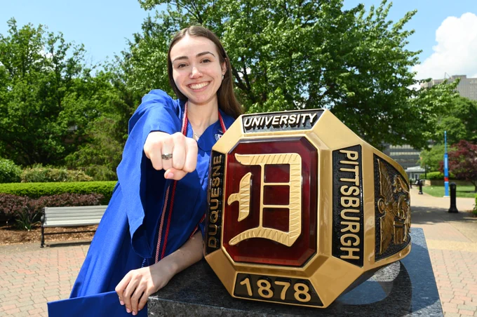 A Duquesne University senior in graduation regalia stands next to the Red Ring statue.