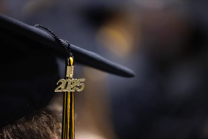 Graduates in caps and gowns, some with honor cords, smiling during a commencement ceremony.
