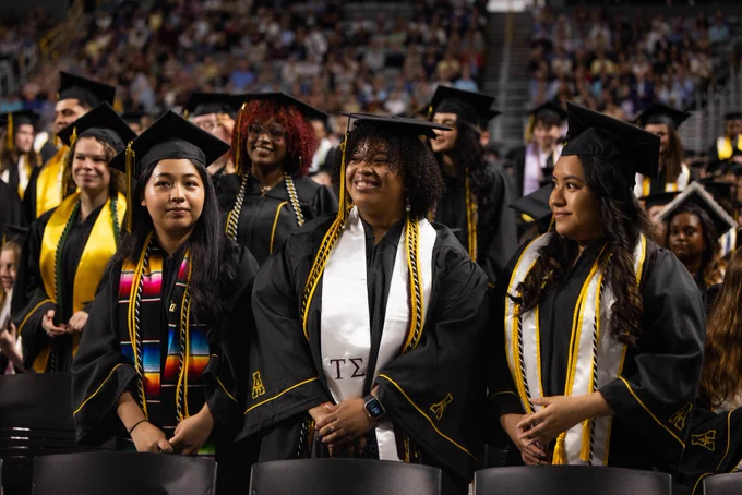 Group of graduates in caps and gowns, smiling and standing together at a commencement ceremony.
