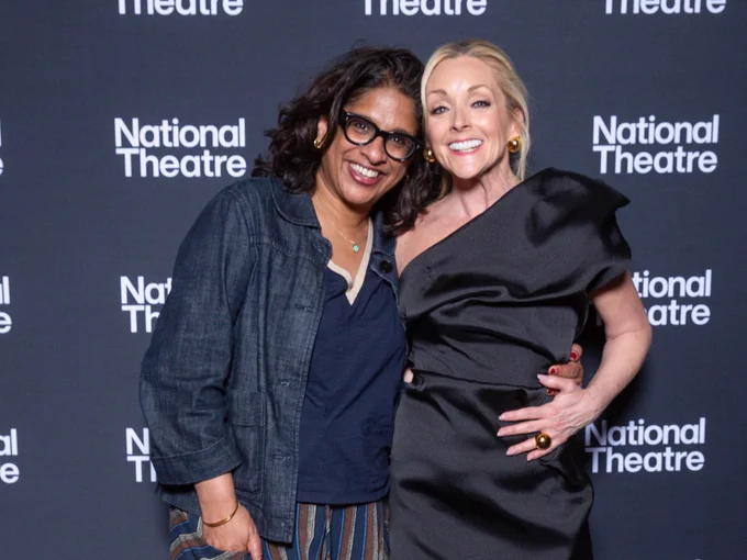 Two individuals smiling and posing together at a National Theatre event, one dressed in a denim jacket and striped trousers, the other in an elegant black dress.