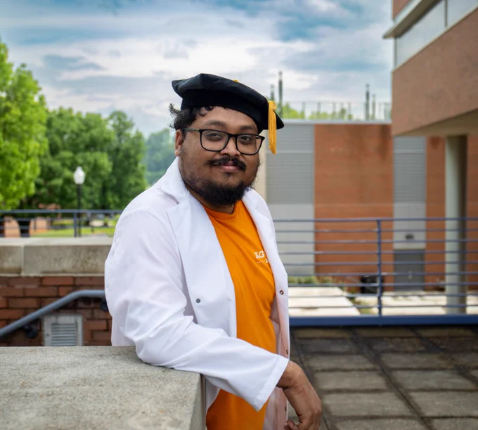 A graduate wearing a white lab coat and a black graduation cap with a gold tassel stands confidently on a campus with buildings in the background.