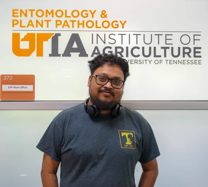 Person standing in front of the University of Tennessee Institute of Agriculture sign, wearing a blue t-shirt with a logo and headphones around neck.
