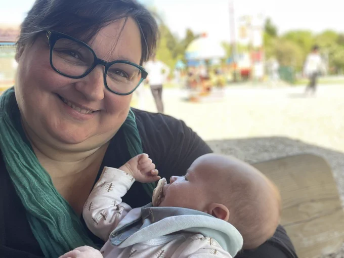 A middle-aged woman wearing glasses smiles while holding a newborn child in her arms