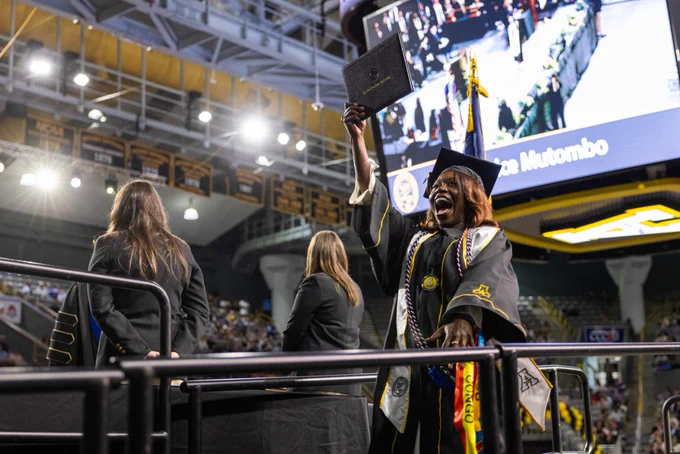 Graduate joyfully holding up diploma at Appalachian State University's commencement ceremony, large screen displaying event in background.

