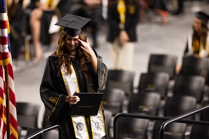 A graduate in academic regalia holds a diploma while walking past rows of empty seats at a graduation ceremony, wiping a tear from her eye, with an American flag visible to the side.
