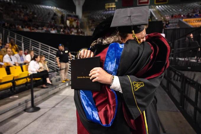 Two graduates hugging at a commencement ceremony. One is holding a diploma folder from Appalachian State University.
