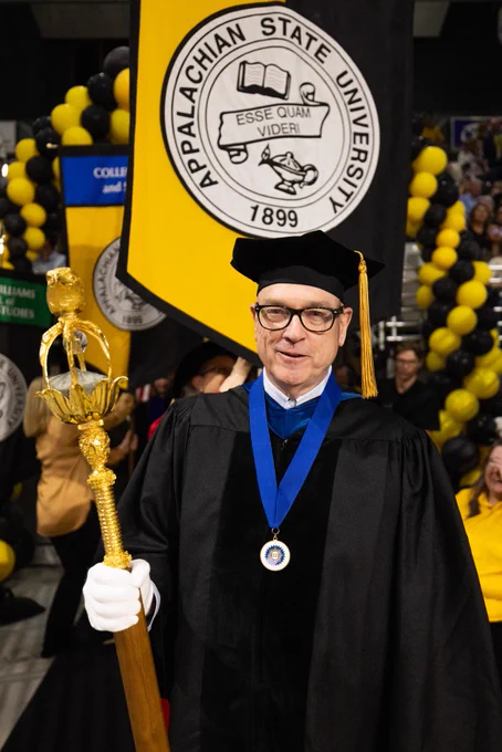Person in academic regalia holding a ceremonial mace at an Appalachian State University graduation ceremony, surrounded by yellow and black balloons.
