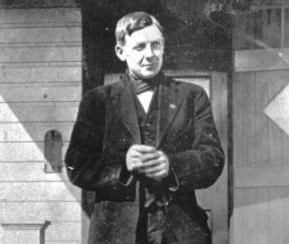 A person stands in formal attire, including a suit and bow tie, in front of a wooden building with a door. The black-and-white photo has "Ginger Goowin" written at the bottom. The style suggests it’s from the late 19th or early 20th century.