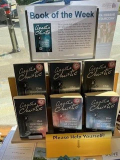 Image is a photograph of Horsley Library's Book of the Week display, a plastic stand featuring 6 copies of Agatha Christie's "The Murder of Roger Ackroyd". Above the display is a laminated advertisement in landscape format. Said advertisement is titled: 'Book of the Week' and has a picture of the Book's cover on the left and the text of the blurb on the right, as in the body of the post.