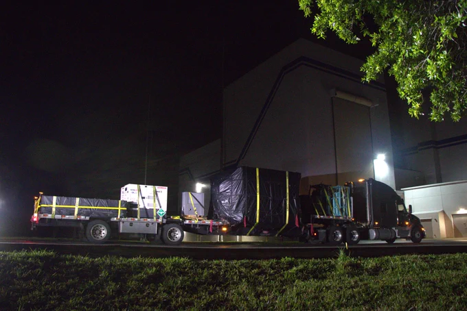A semitractor-trailer pulls a long, multi-axle flatbed trailer carrying a large white metal box that contains NASA's Interstellar Mapping and Acceleration Probe or IMAP spacecraft. It is in front of a tall, white industrial building with a large roll-up door. 