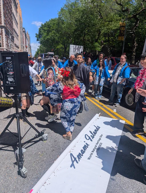 A group of people dressed in traditional Japanese clothing parading down a sunny street, with some dancing and others watching. In the foreground is a rolled-up banner reading “Amami Islands – World Natural Heritage Site UNESCO.”