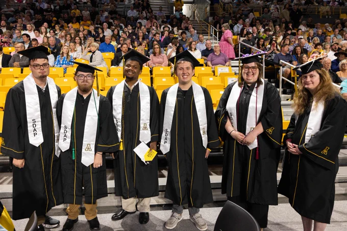 Five graduates in black caps and gowns with honor cords, holding their diplomas at a university graduation ceremony, with an audience in the background.