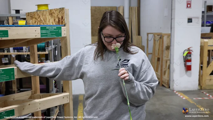 A young woman with glasses looks down while smelling a peach colored carnation. Her hand is resting on a wooden label cart. The bottom left hand corner has text that reads, “LayerZero® is a registered trademark of LayerZero Power Systems, Inc. All rights reserved.” The bottom right hand corner has the official logo and below it reads the website name, “www.LayerZero.com”.