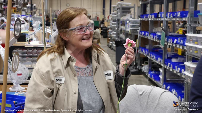 An older woman with glasses stares off screen with a smile while holding a pink carnation. She is wearing a jacket that reads “Sheila” on the left and the LayerZero logo on the right. Behind her is the wiring area. The bottom left hand corner has text that reads, “LayerZero® is a registered trademark of LayerZero Power Systems, Inc. All rights reserved.” The bottom right hand corner has the official logo and below it reads the website name, “www.LayerZero.com”.