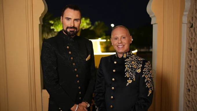 Rylan and Rob dress in black with gold trim while attending Diwali celebrations at the Jaipur City Palace. They stand in an arch at night, smiling.