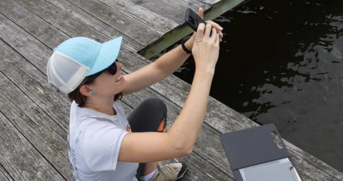 A woman on a pier points her phone up to the sky while using the HydroColor app to collect water clarity data