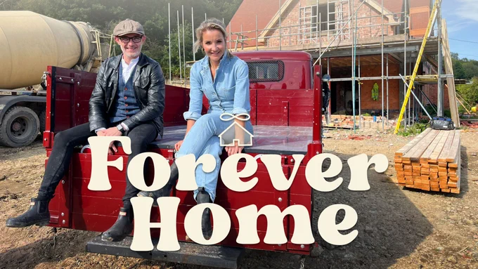 A woman and man are sitting on the back of a pickup truck smiling towards the camera. In the bacground we can see a house with scaffolding around it and there is a cement mixer behind the truck. 