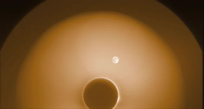 A bright, circular fuzzy halo surrounds the Sun, blocked by a dark disk in the center of the image. Within the halo, a small, detailed image of the Moon appears off-center against a brownish background, creating a cosmic, ethereal effect.

