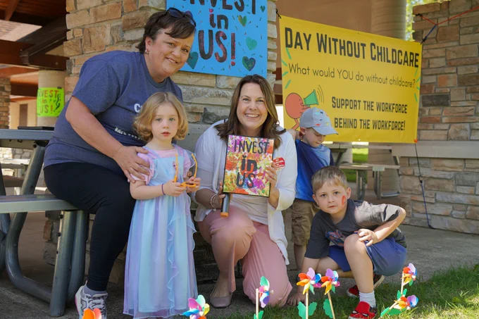State Senator Dianne Hesselbein kneels to take a photo with an advocate and a group of children at the Day Without Childcare event in Waunakee. Senator Hesselbein is smiling and holding a painting made with colorful handprints written over in black text that says "invest in us." Behind them is a yellow banner proclaiming the Day Without Child Care.