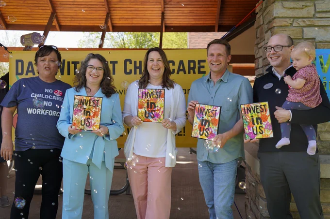 State Senator Dianne Hesselbein poses for a group photo with County Executive Melissa Agard, State Representative Alex Joers, and DCF Secretary Jeff Pertl. They are also joined by Heather Murray, an advocate and preschool owner. Each of the elected officials are holding a painting with colorful handprints and black text that reads "invest in us." There are also small bubbles floating in the air, slightly distracting everyone and causing them to laugh.