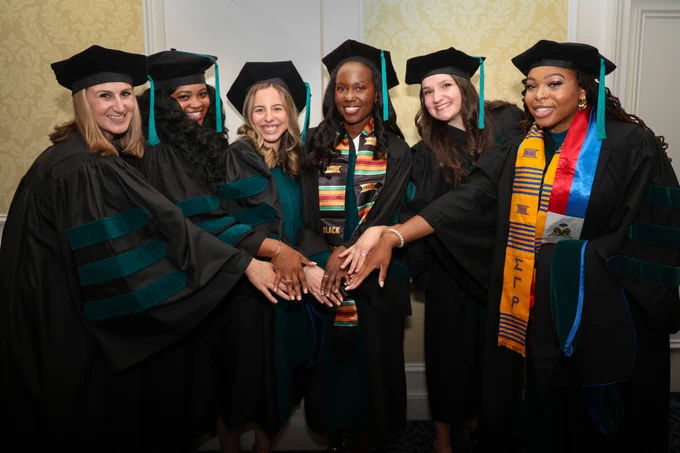 Six graduates in academic regalia smiling and holding hands, showcasing various stoles and cords at their graduation ceremony.