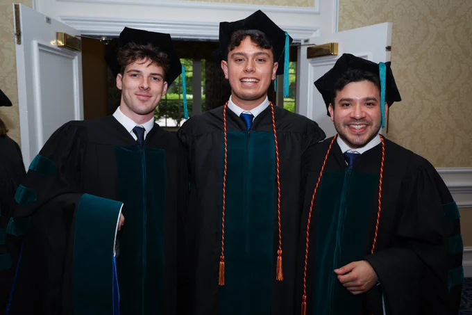 Three graduates in green and black gowns smiling at a graduation ceremony.
