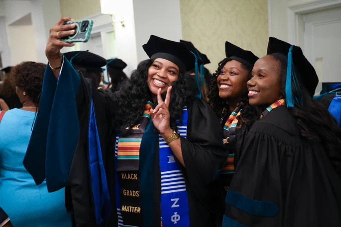 Three graduates in caps and gowns taking a selfie, one making a peace sign, with a stole that reads "Black Grads Matter.
