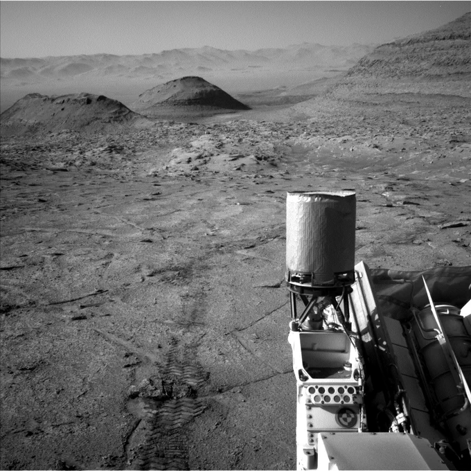A black and white image taken by the Curiosity rover shows a rocky, desert-like landscape with wheel tracks in the foreground. A cylindrical instrument is mounted on the rover’s deck in the bottom right corner. In the distance, several rounded hills rise from the flat terrain, and layered rock formations line the right edge. The hazy Martian horizon stretches across the background.