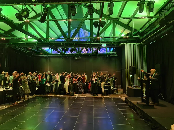 A man on a stage on the right with an audience on the left holding up wine glasses in a toast. 