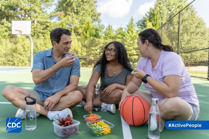A father, mother, and daughter take a break from playing basketball to have a snack of fresh fruit and drink water. 