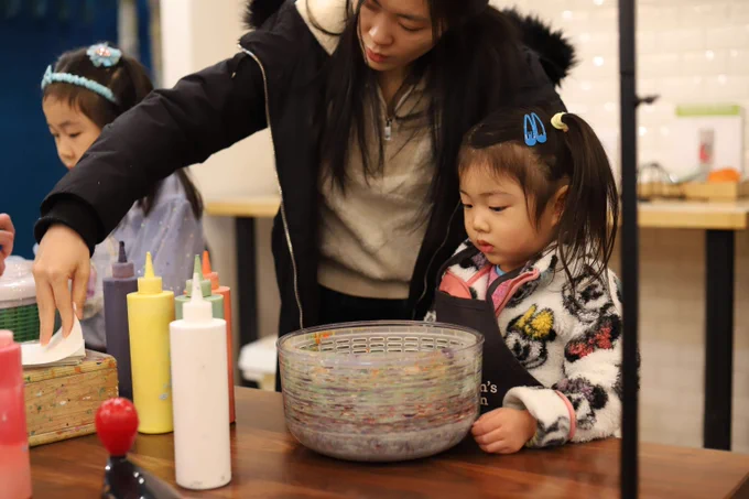 A woman in a black winter coat with fur trim helps two young girls with an art project. The girls, both with hair accessories, are focused on a colorful bowl-shaped container on the wooden table. Various paint bottles and art supplies surround them. The setting appears to be an arts workshop space.