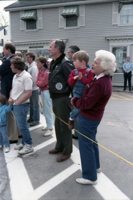 A group of people, including Barbara Bush holding a child and George H.W. Bush wearing a leather jacket with a patch, standing by a street observing an event.